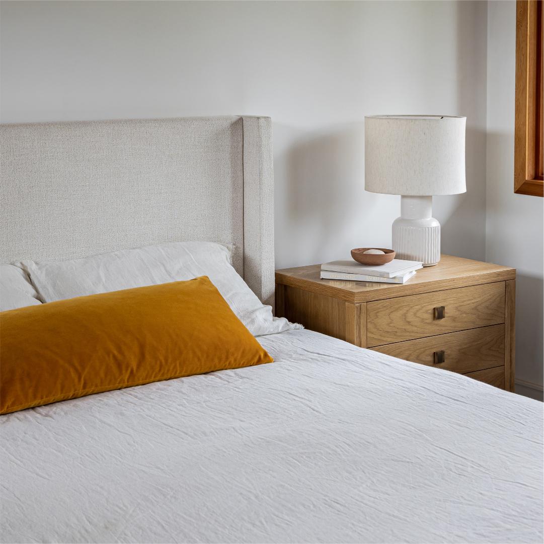 Bedroom with a bed featuring a mustard yellow pillow, a wooden nightstand with a lamp, and books.