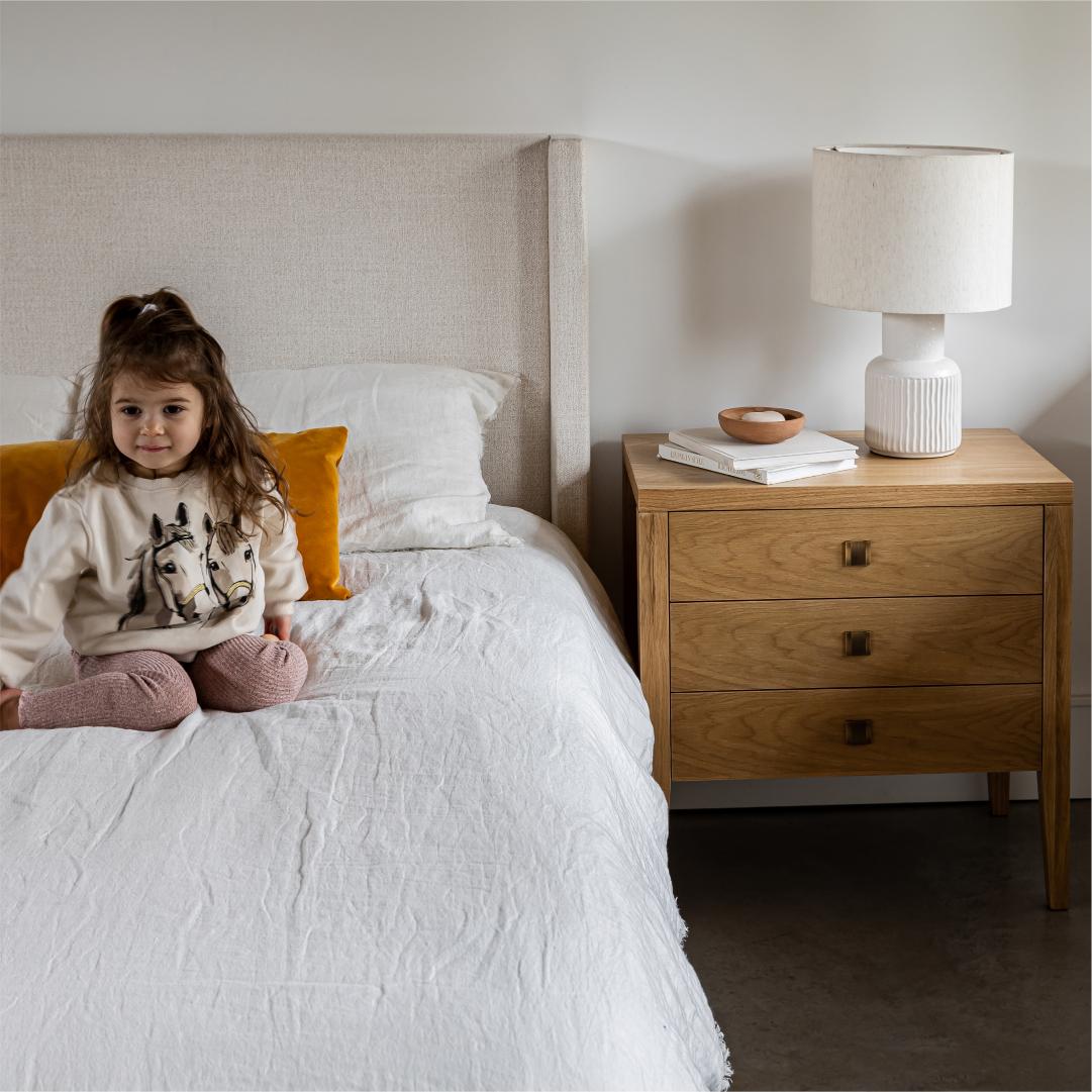 Child sitting on a bed in a bedroom with a wooden nightstand and lamp.