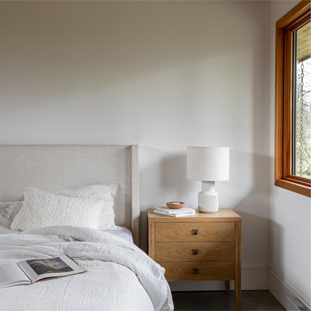 Bedroom with wooden nightstand, lamp, and open book near a window.