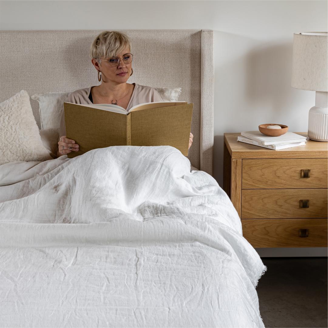 Woman reading a book in bed with a wooden nightstand next to her.