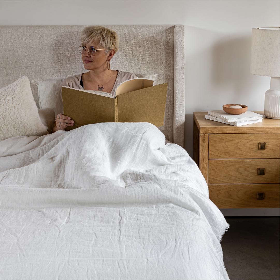 Woman reading a book in bed with a wooden nightstand next to her.