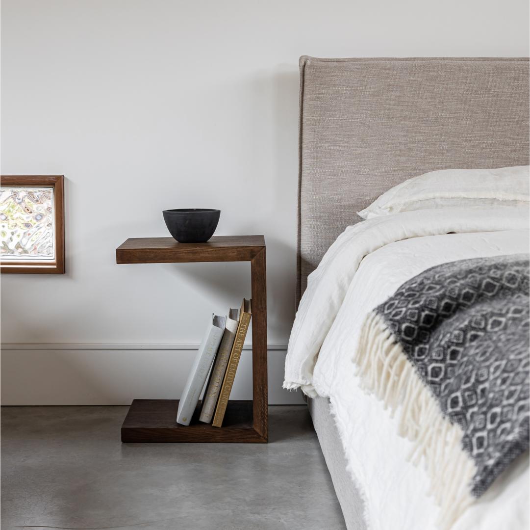 Bedroom with a wooden side table, books, and a bowl next to a bed with white bedding and a patterned throw.
