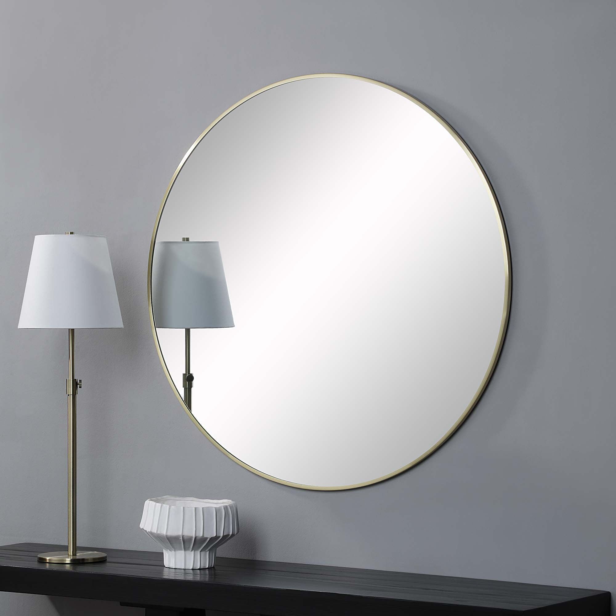 Grady round mirror with a gold metal frame, reflecting a white lamp and a textured white vase on a dark wooden table.