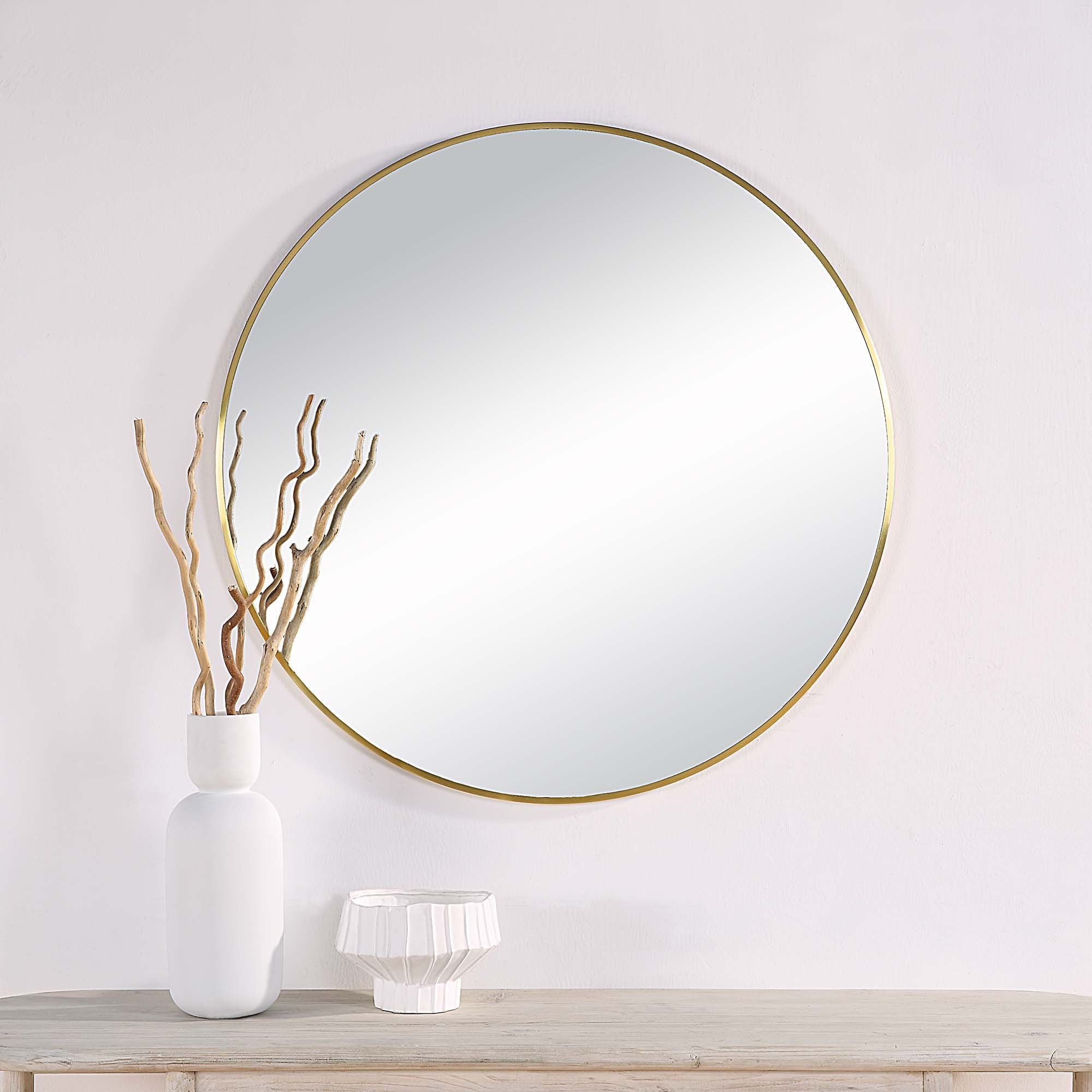 Parga round mirror with a thin gold frame, reflecting light, displayed above a wooden table with white vases and decorative branches.