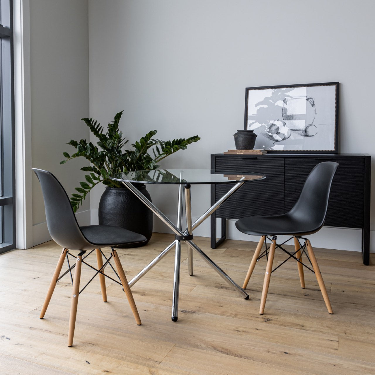 Modern glass dining table with black chairs in a room with a plant and framed picture.