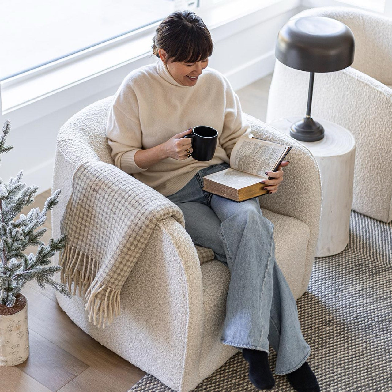 Person sitting in a cozy armchair reading a book and holding a mug.