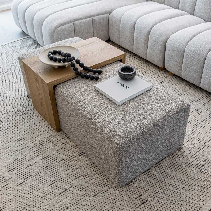 Gray textured ottoman with a wooden side table and decorative items in a living room setting.