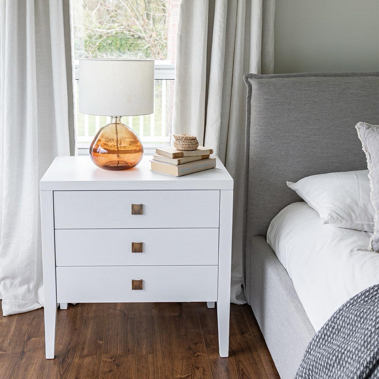 White nightstand with a lamp and books next to a bed in a bedroom.