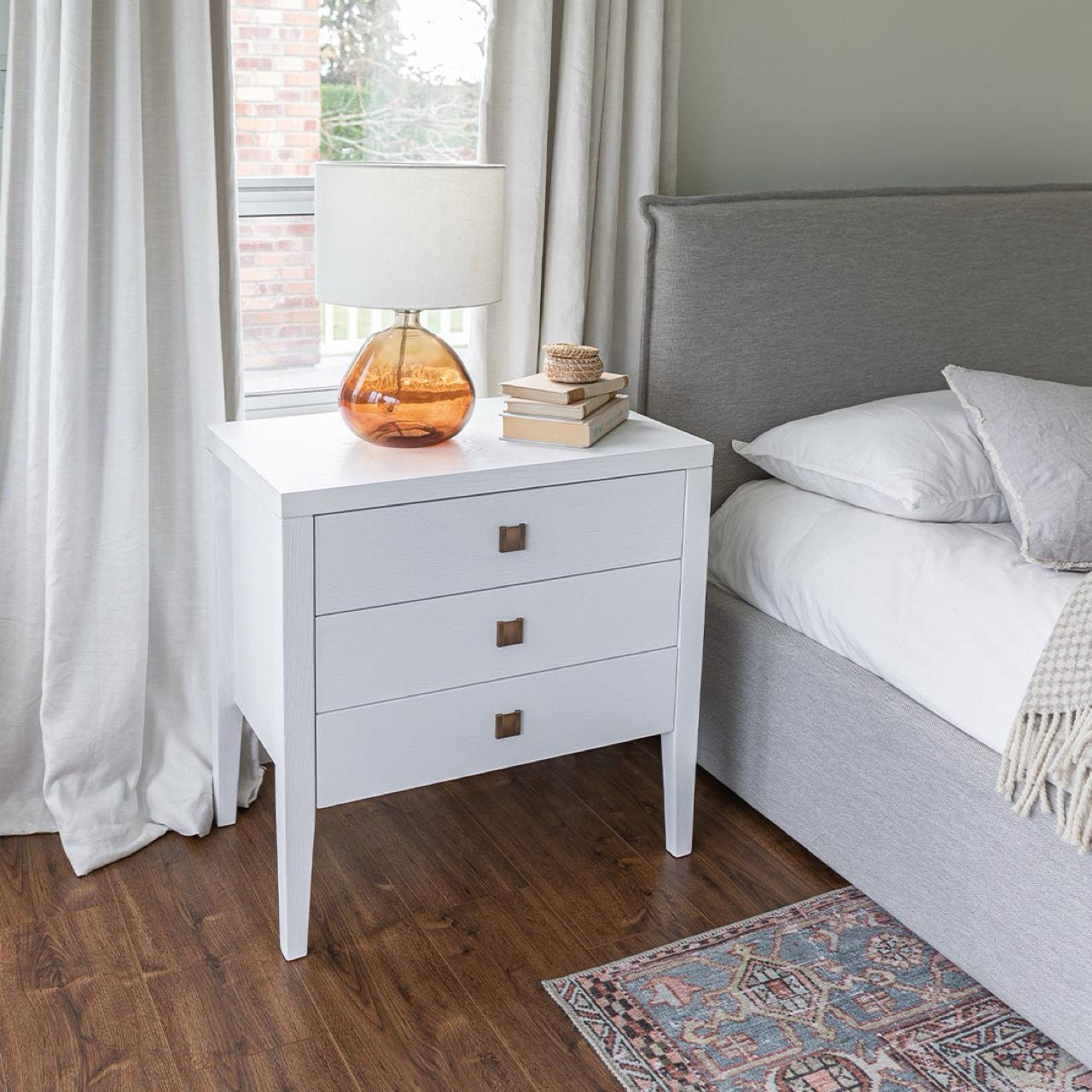 White nightstand with lamp and books next to a gray upholstered bed in a bedroom.
