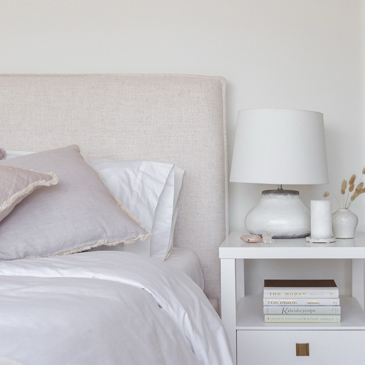 Neatly made bed with pillows and a nightstand with a lamp and books in a bedroom setting.