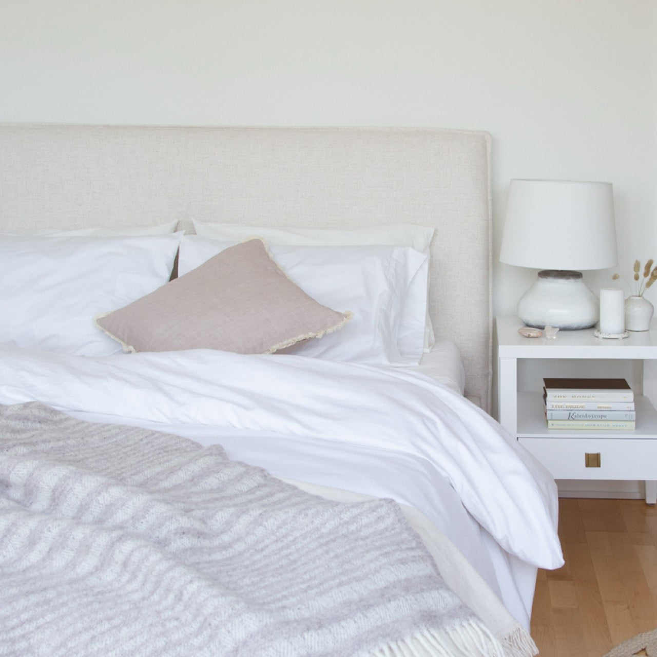 Neatly made bed with white bedding and a beige pillow, nightstand with lamp and books in a bedroom.