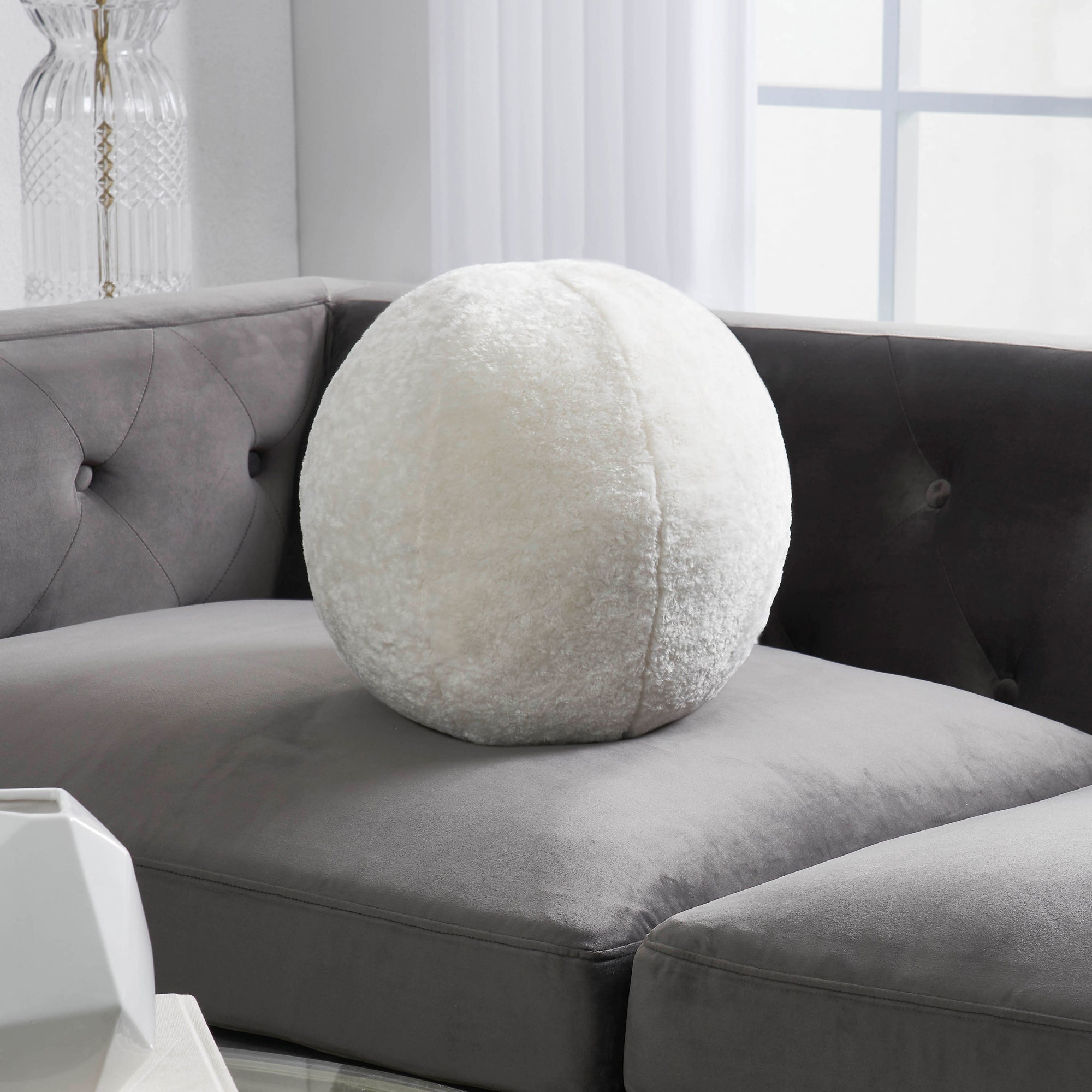 Freddie Indoor Pillow placed on a gray couch, showcasing its round shape and fluffy cream fabric.