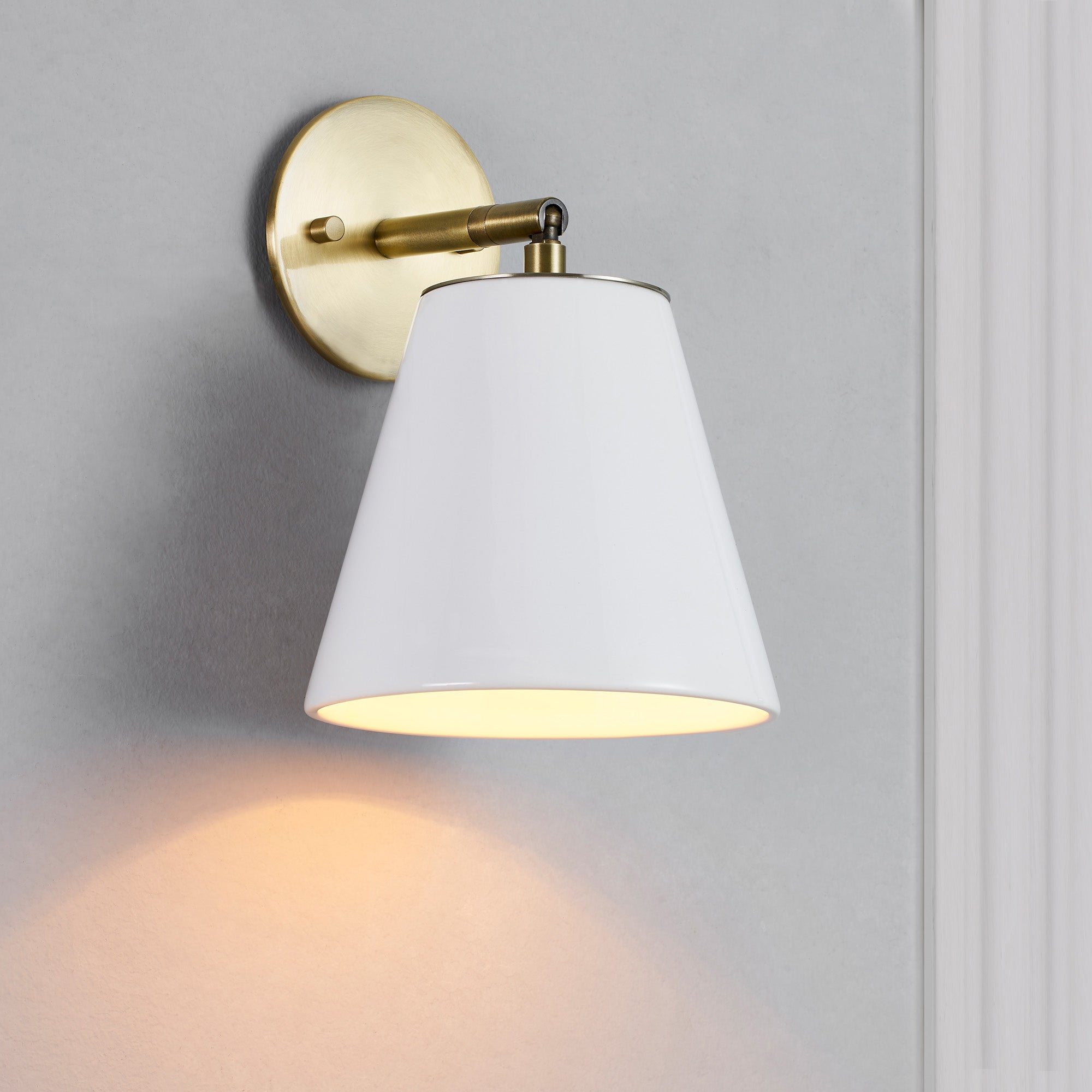 Kai Wall Sconce featuring a white conical shade and a brass wall mount, casting warm light on a gray background.