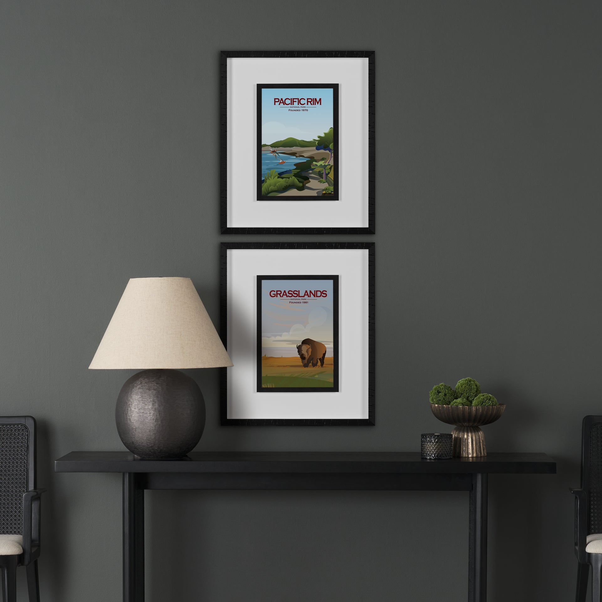 Two framed posters on a dark wall above a black console table with a lamp and plant.