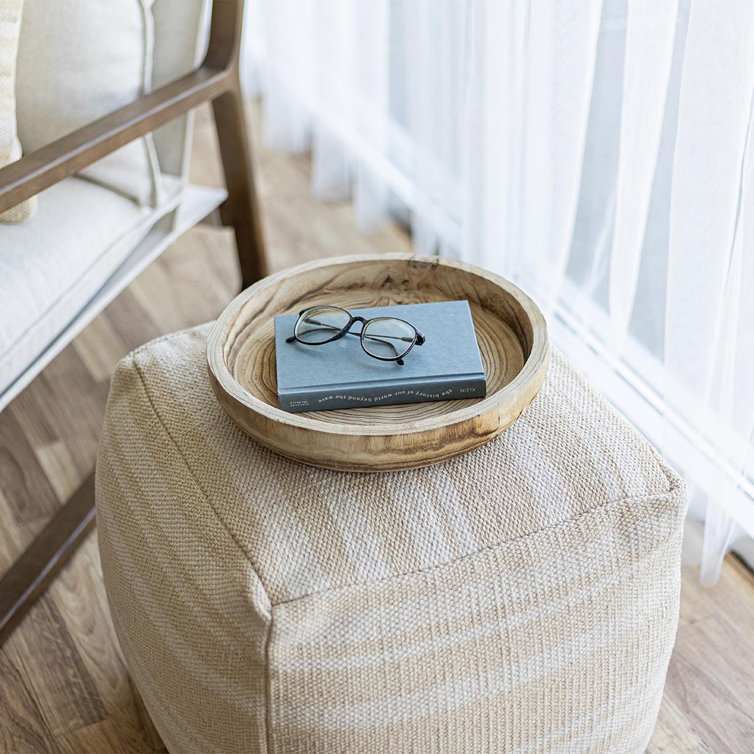 Wooden tray with a book and glasses on a textured ottoman in a room with light filtering through sheer curtains.