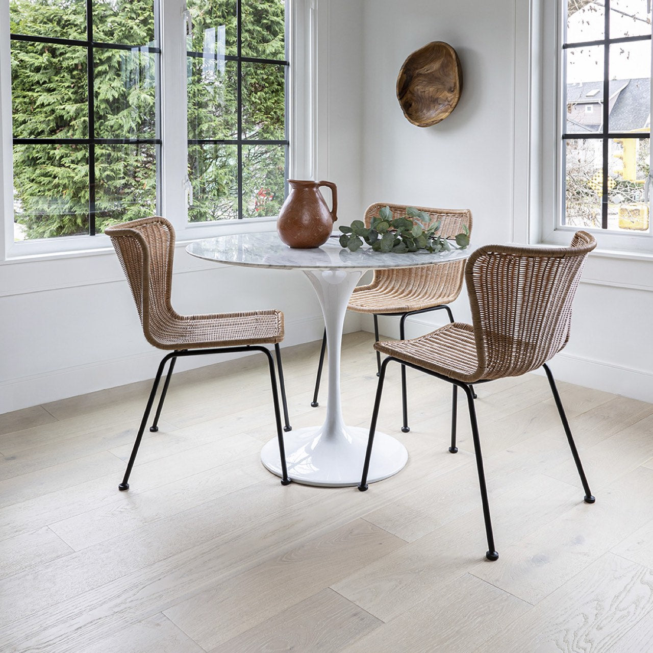 Dining area with round table and wicker chairs in a bright room with large windows.