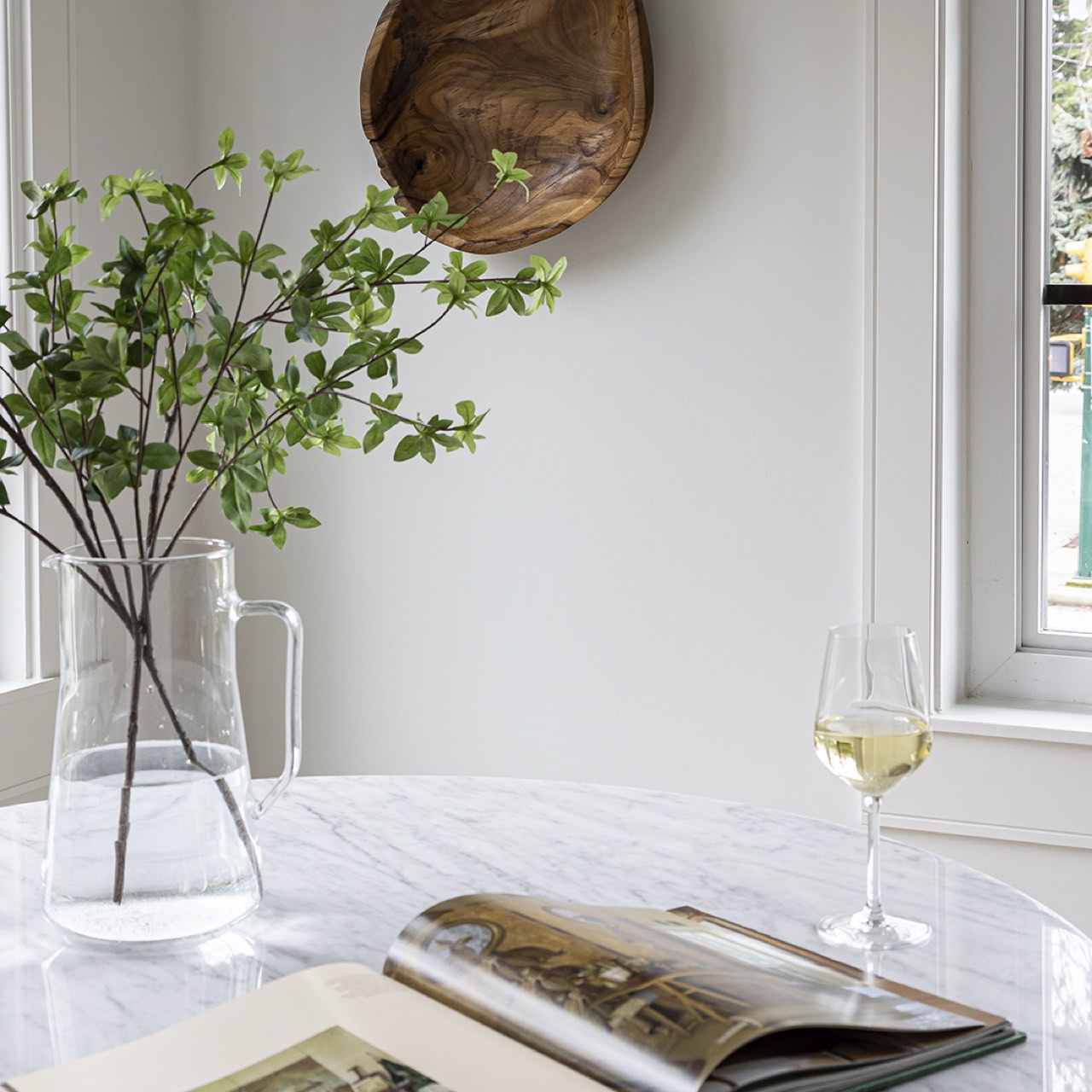 Clear vase with greenery, open book, and glass of white wine on a marble surface.