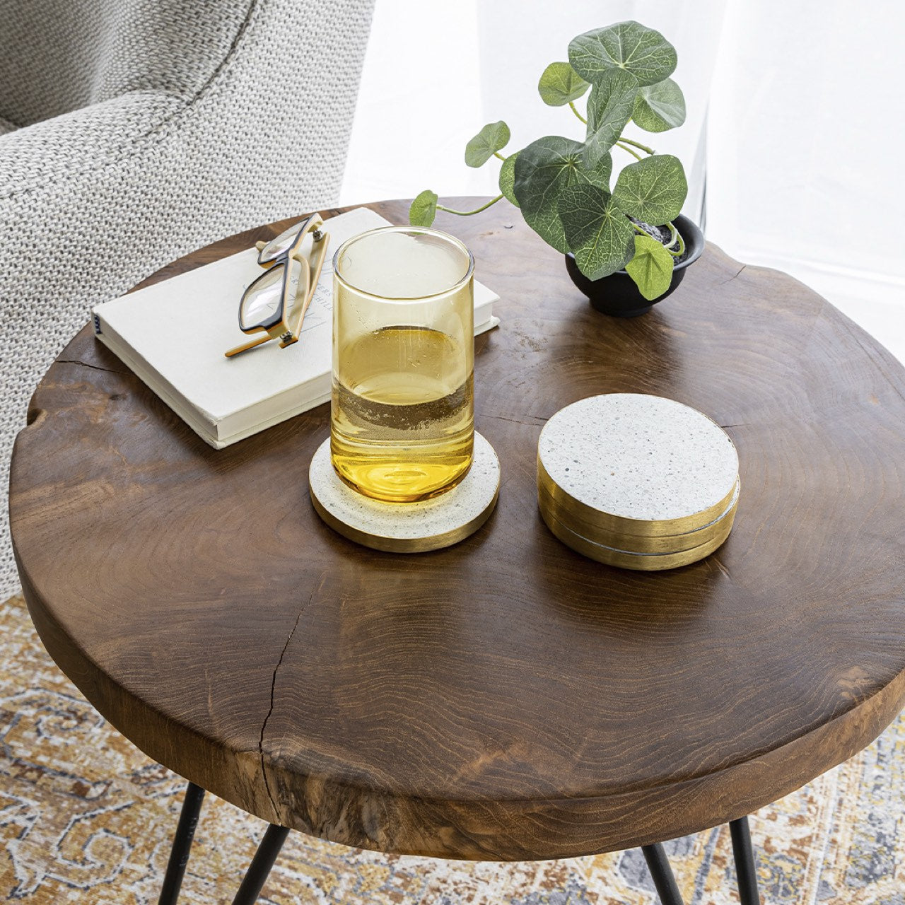 Round wooden table with a glass of yellow liquid, coasters, and a plant in a cozy indoor setting.
