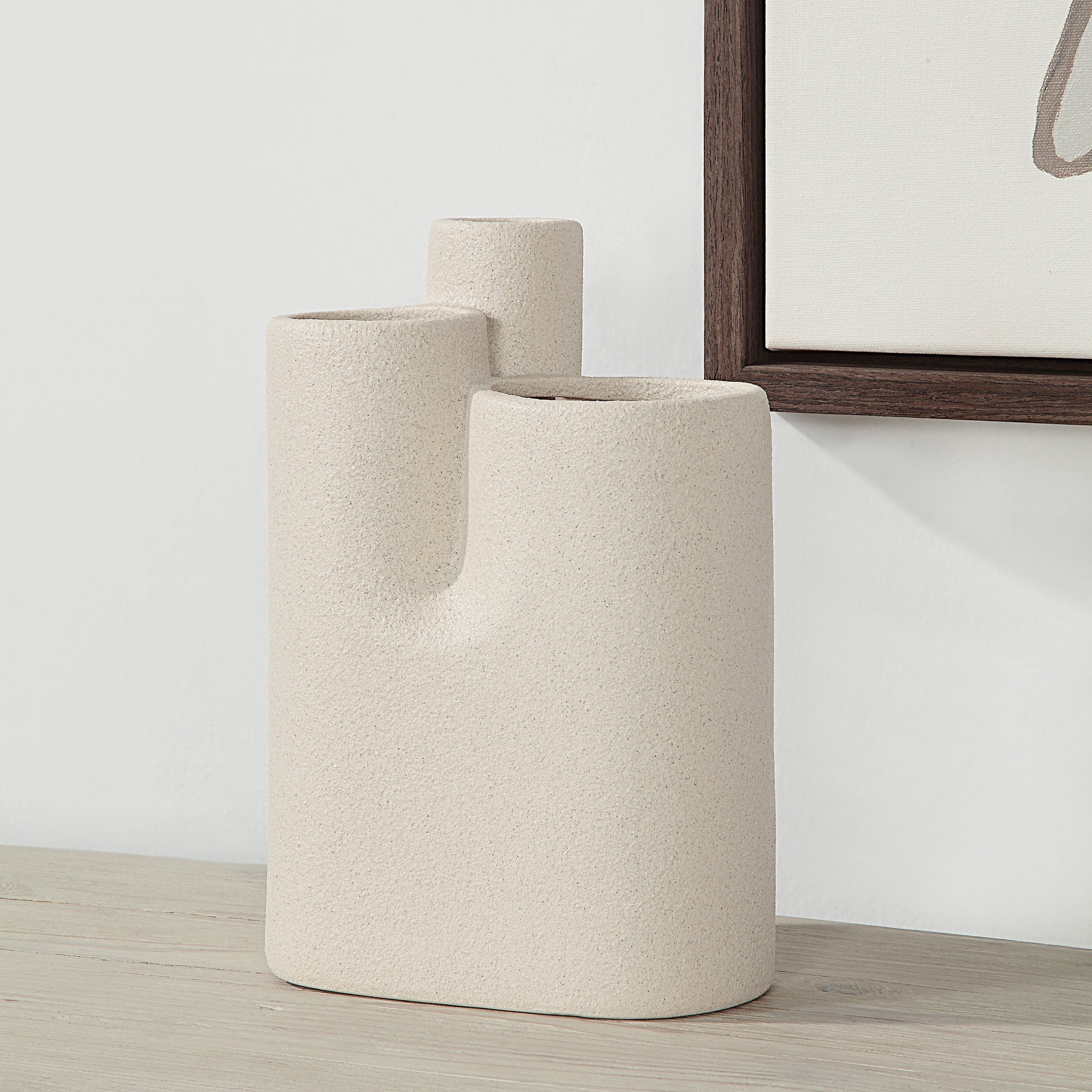 Fontaine Vase by Renwil in beige ceramic, displayed on a wooden surface next to artwork.
