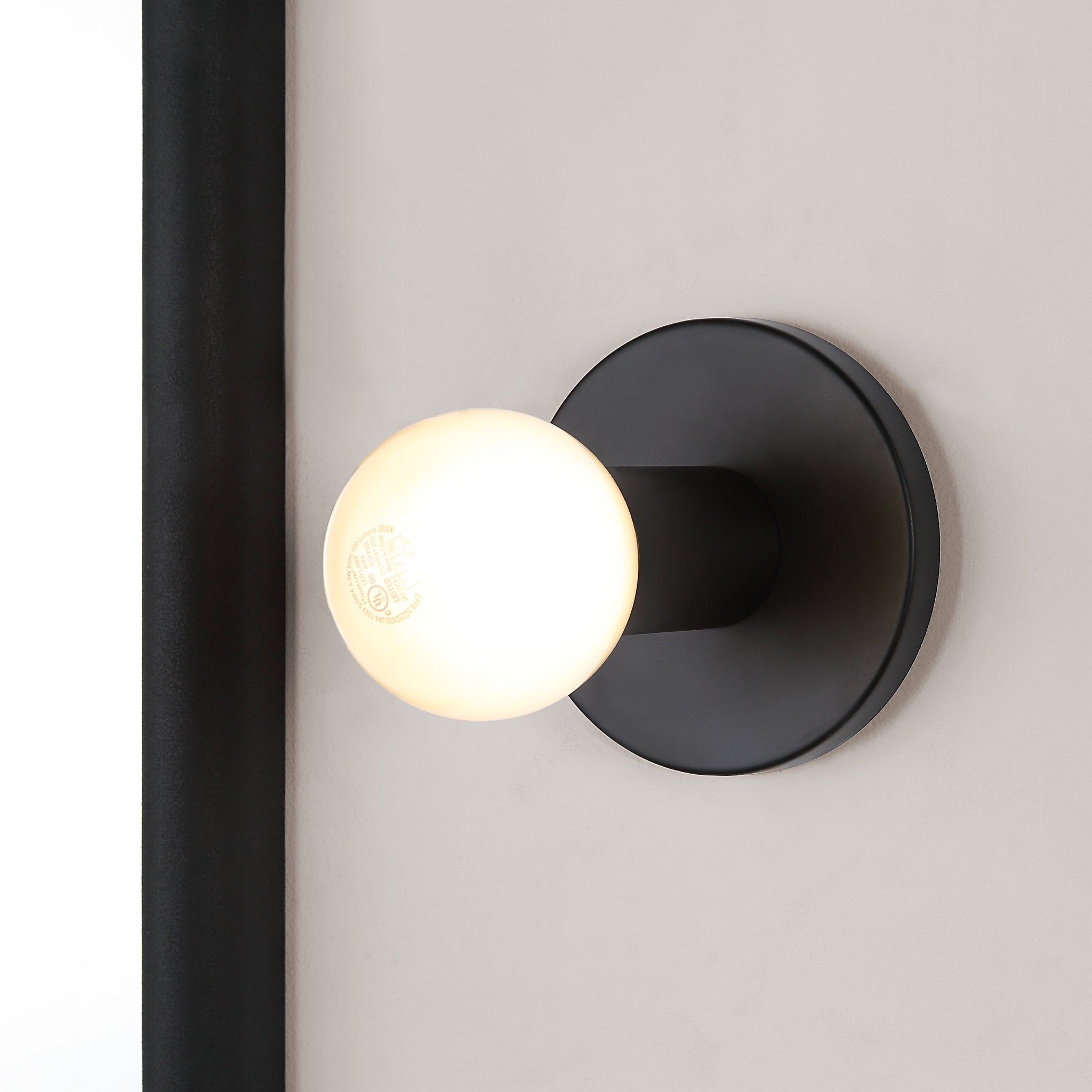 Thessa Wall Sconce installed on a wall, showcasing black mount and glowing white bulb.