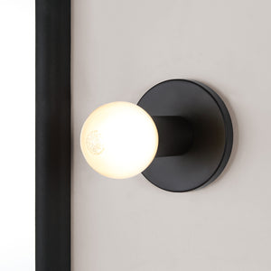 Thessa Wall Sconce installed on a wall, showcasing black mount and glowing white bulb.