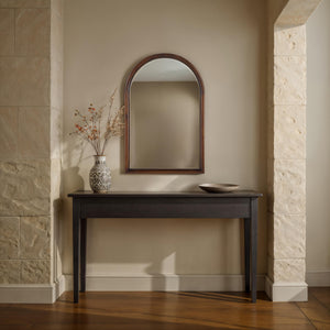 Lanai mirror with an arched wooden frame above a dark console table, featuring a decorative vase and bowl.