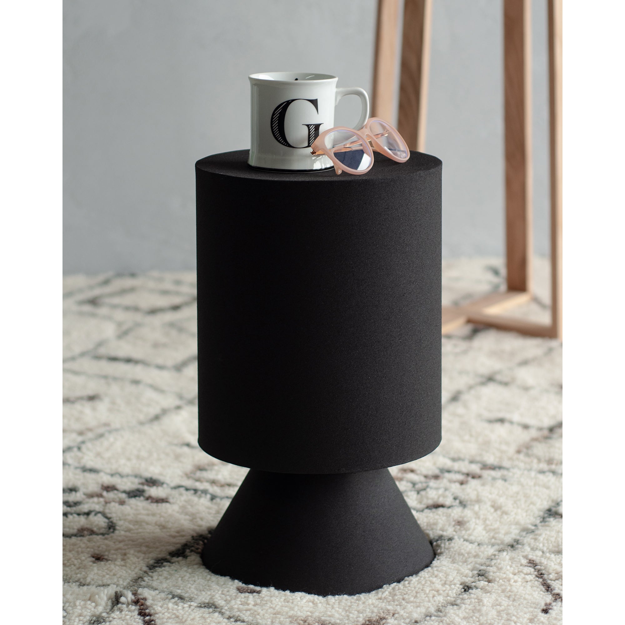 Oloria Stool in black with a coffee mug and sunglasses on top, placed on a patterned rug.