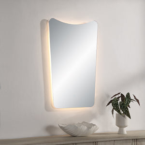 Rimini Led Mirror mounted on a wall, illuminated with soft lighting, beside a plant.